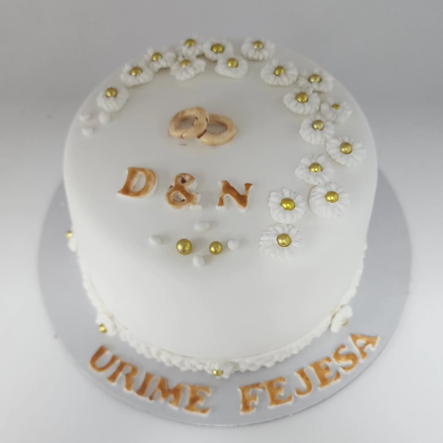 Torte e bardhë fejese me lule të bardha dhe unaza të arta